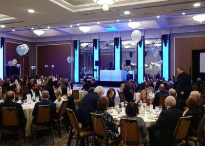 Elegant dinner event with guests seated at round tables, a speaker at the podium, and decorative blue and white balloons.