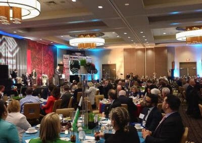 A corporate event in a banquet hall, with attendees seated at tables listening to a speaker at a podium.