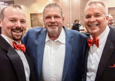 Three smiling men at a formal event, two wearing black tuxedos with red bow ties and one in a suit without a tie.