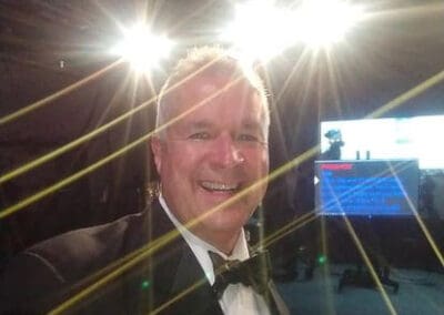 A man in a tuxedo smiling at the camera, with bright stage lights creating a flare effect behind him.