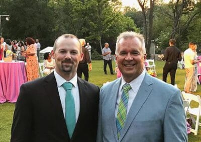 Two smiling men in suits—one with a teal tie, the other with a green pattern—stand outdoors amid guests and trees.