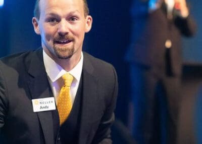 Man in a black suit and yellow tie smiling at an event,; his badge reads 'Andy'. Another man is speaking in the background.
