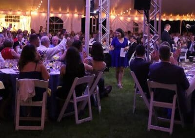 A woman in a blue dress walks among tables at an evening event under a tent with string lights and seated guests.