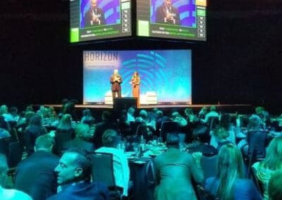 Two presenters on stage at a fundraiser with guests seated at round tables, looking at a large screen displaying the presenters.