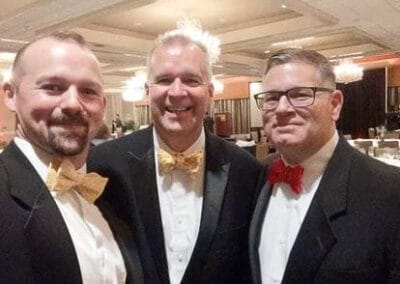 Three smiling men in tuxedos with bow ties at a formal event.
