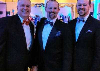 Three men in tuxedos, each wearing bow ties, smiling at a formal event with a brightly lit, decorated background.
