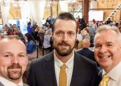 Three men in suits and ties smiling for a selfie at a crowded indoor event with decorations and chandeliers.