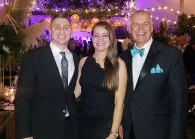 3 people smiling at an evening event: two men in suits, a woman in a black dress, with colorful lights and trees in background.
