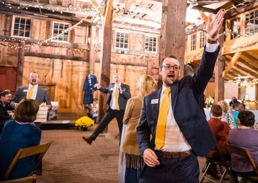 A man in a suit raises his hand at an auction with guests seated around tables, in a rustic venue decorated with string lights.