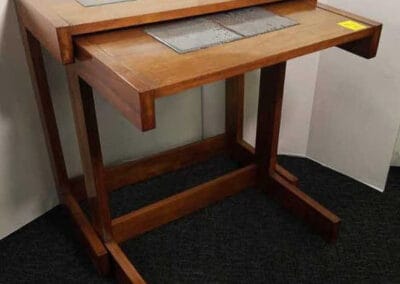 A wooden drafting table with angled top and built-in rulers, on a dark carpeted floor against a white wall.