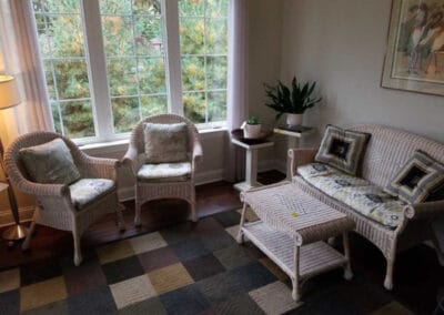 Living room with wicker furniture, with two chairs a sofa with cushions, a floor lamp, and large windows overlooking greenery.