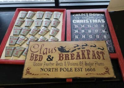 A wooden Christmas countdown calendar next to a sign reading "Claus Bed & Breakfast, North Pole est 1605" with holiday motifs.