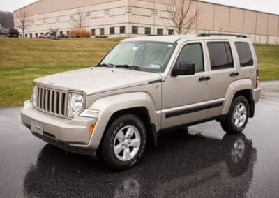 A light colored Jeep parked on a wet road with an industrial building in the background.