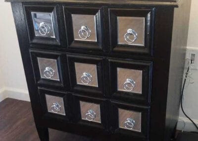 A small black wooden cabinet with nine shiny, silver ring handles on its drawers, against a light-colored wall.