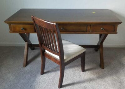 A wooden writing desk with two drawers with a wooden chair featuring a white cushioned seat.