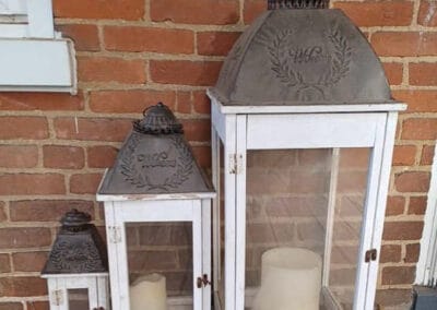 Three rustic lanterns of varying sizes, each with a metal top and a white wooden frame, displayed against a red brick wall.