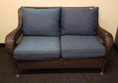 A two-seater, wicker couch with blue cushions against a plain wall in an indoor setting.