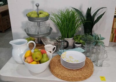 Kitchenware display with pitchers, mugs, plates, a three-tiered fruit stand, assorted fruits, and artificial plants on a table.