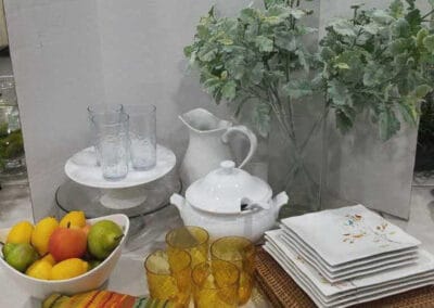 A kitchen display featuring a bowl of fruit, amber glasses, a white teapot, and plates, with a potted plant in the background.