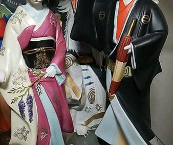 Two traditional Japanese ceramic figurines, a woman in a pink kimono and a man in a black and white ensemble, on a shelf.