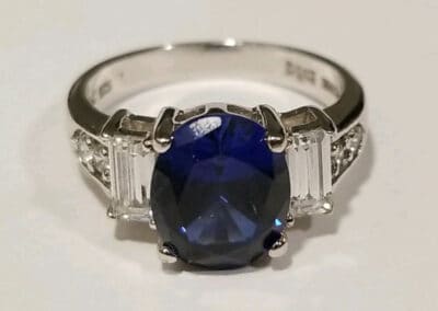 A silver ring with a large blue sapphire, flanked by two smaller rectangular diamonds on a white background.
