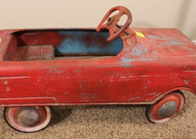 Vintage red pedal car with noticeable wear and chipped paint, displayed on a carpeted floor.