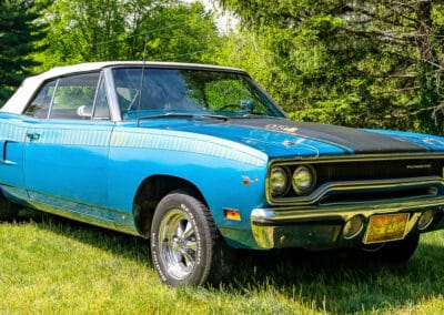 A vintage blue Plymouth Roadrunner car parked on the grass, showcasing its classic design and gleaming chrome details.
