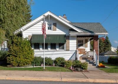 A quaint white single-story house with a green awning, American flag, and front porch, under clear skies on a sunny day.