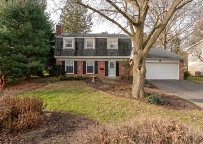 Two-story suburban house with white siding, black shutters, attached garage, and a landscaped yard with a large tree.