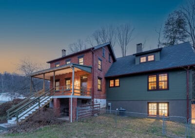 Two-story red brick farm house with a green siding addition, lit windows, and a porch, located on a hillside during twilight.