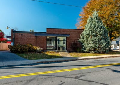 A small brick office building with a flat roof and glass front door, flanked by colorful trees, located beside a street.