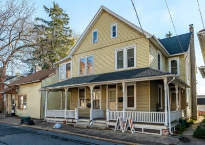 Two-story yellow house with a covered porch, white railings, and a clear blue sky, in a suburban neighborhood.