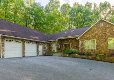 A one story stone house with a two car garage, surrounded by lush greenery under a clear sky.