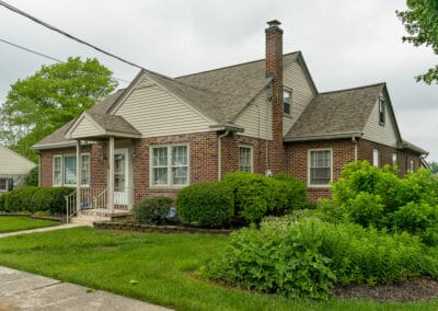 A brick suburban house surrounded by lush green bushes and a well-maintained lawn, under an overcast sky.