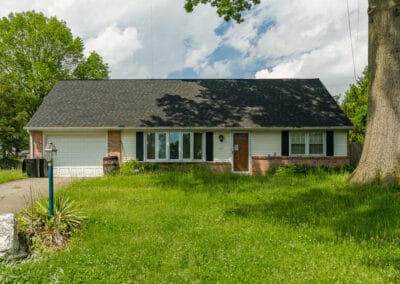 A one-story brick house with white siding under a large tree, featuring an overgrown lawn and a partly cloudy sky.