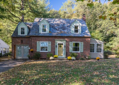 A charming two-story brick house surrounded by trees and a manicured lawn, with mums near the entrance.