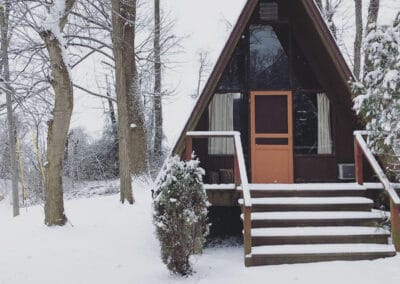 A small A-frame cabin with an orange door, surrounded by snowy trees and ground.