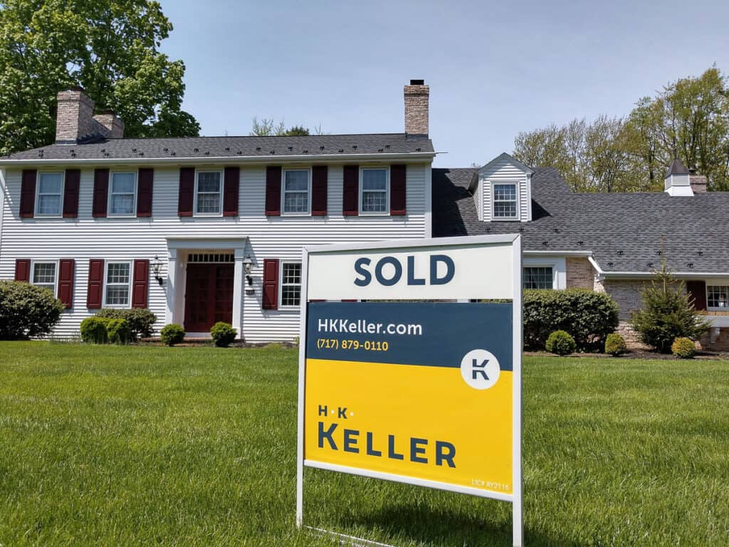 "Sold" sign from H. K. Keller in front of a two-story house with red shutters and white siding under a clear blue sky.