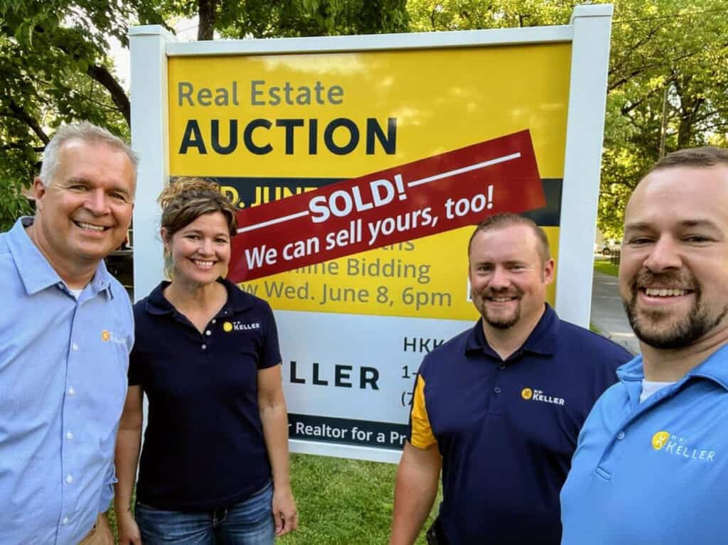 Four people smiling in front of a real estate auction sign that announces a property sold.