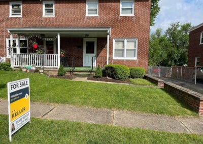 Brick duplex with an H. K. Keller "for sale" sign in the front yard, featuring a porch and well-kept lawn under a clear sky.