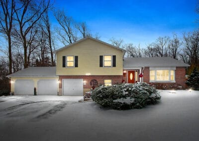 A split-level home with a three-car garage, surrounded by snow-covered trees and bushes, during twilight.