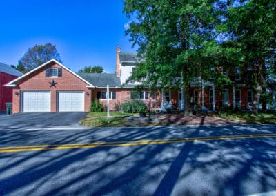 A two-story red brick house, viewed across a road under a clear blue sky, with tree shadows on the pavement.