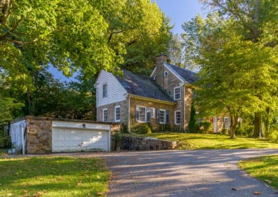 Two-story vintage stone house with white shutters, adjacent garage, and a tree-lined driveway in a sunny, leafy setting.