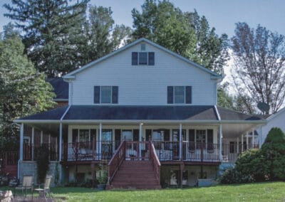 Two-story house with a large front porch and staircase, surrounded by trees and a well-maintained lawn under a clear sky.