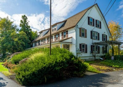 Large white house with a brown roof, surrounded by lush green bushes under a blue sky with clouds.