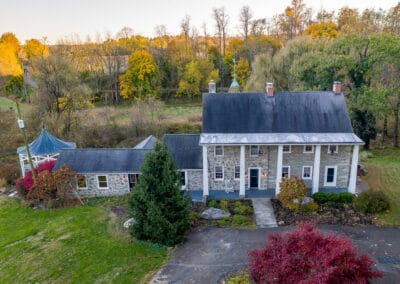 Aerial view of a large, two-story stone house with a grey roof, surrounded by colorful autumn trees and a well-maintained lawn.