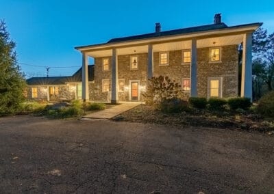 A two-story stone house with outdoor lights at twilight, with multiple windows and surrounded by trees.