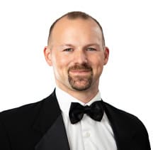 A man with a beard, wearing a gray suit, white shirt, and yellow tie, smiling against a white background.