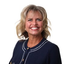 A smiling woman with blonde, wavy hair wearing a white blouse with a grid pattern, on a white background.